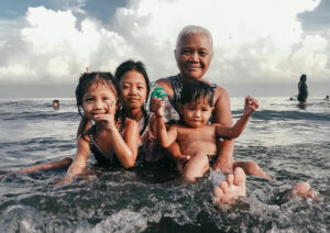Grandmother and three grandchildren sit in shallow ocean water looking to camera.