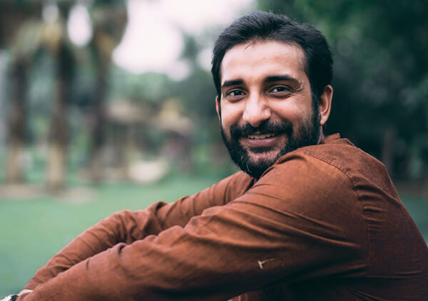 Close up of a smiling man with beard seated on grass outdoors.