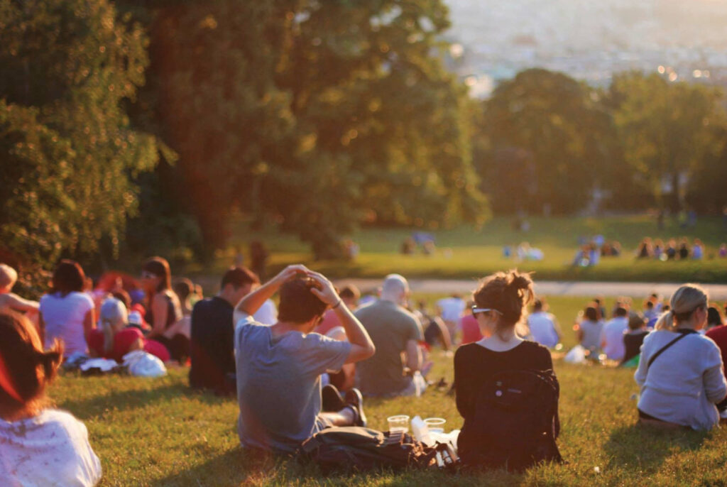 Exterior shot showing a group of people sitting on grass in a park at sunset.