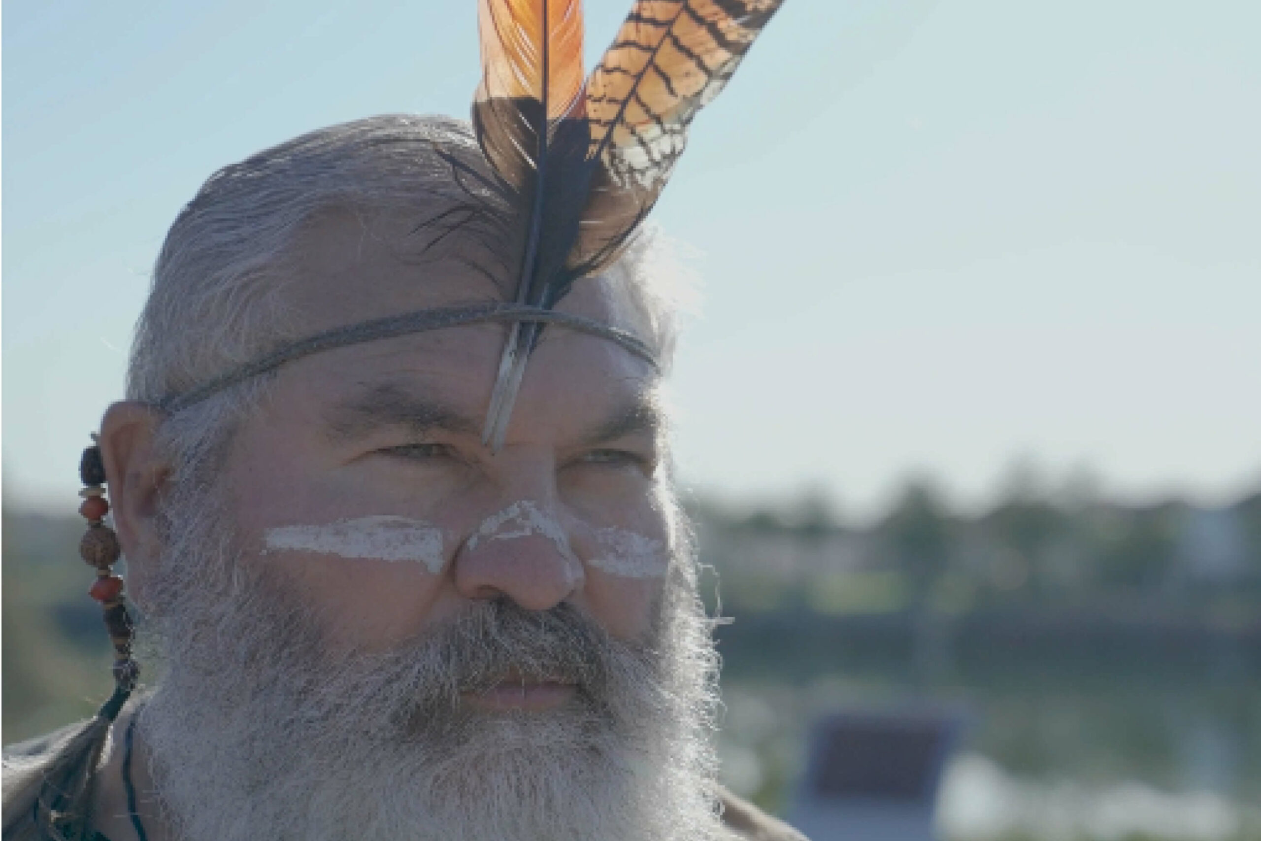 Man in traditional dress with grey beard and feathered headdress, looking into distance.