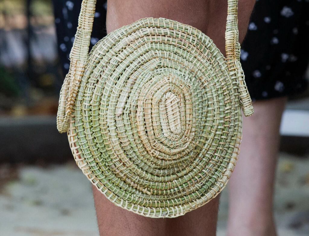 Woven dried grass bag with handle being held in front of legs.