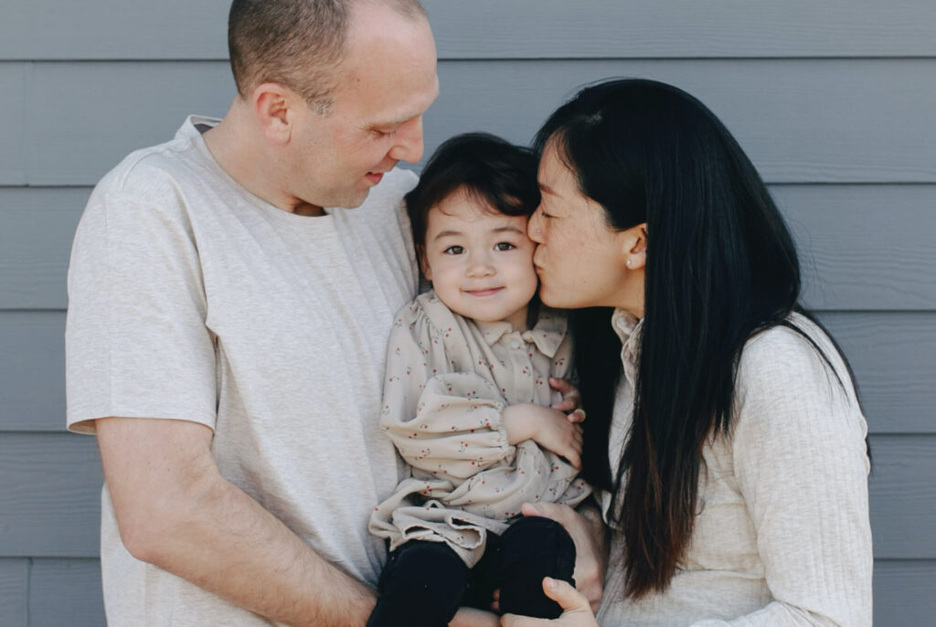 A happy mother and father hold their young daughter, while the mother kisses her on the cheek.