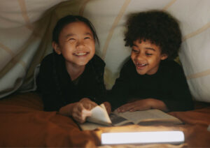 Two kids reading by torchlight under a sheet.